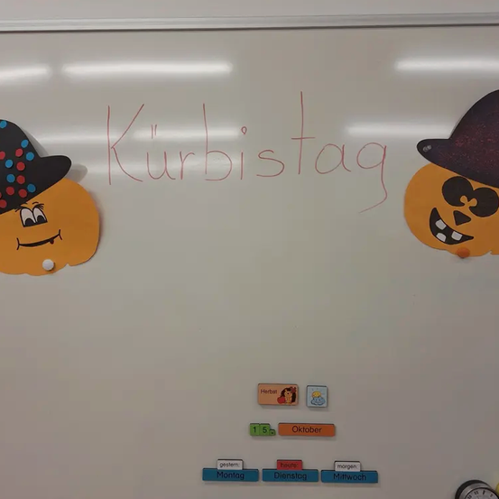 Eine Tafel zeigt 'Kürbistag' in Rot, mit zwei orangefarbenen Kürbissen und einem schwarzen Hut. Darunter befindet sich ein Kalender mit 'Oktober' und Wochentagen.