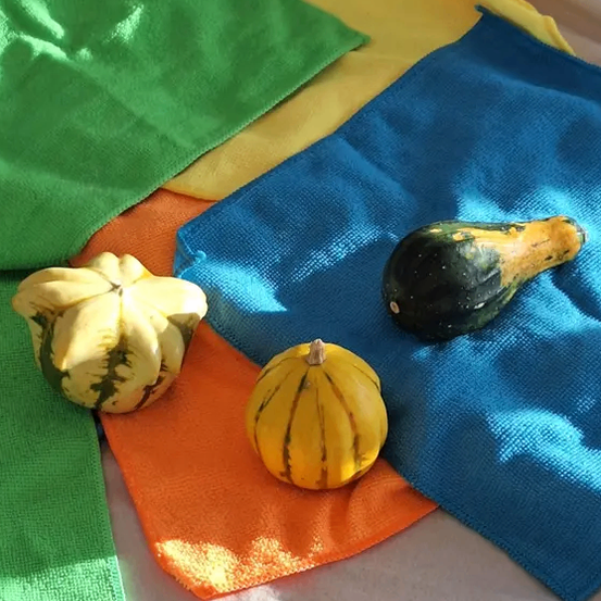 Drei Kürbisse liegen auf einem Bett aus farbenfrohen Tüchern. Ein grünes Tuch ist links, ein gelbes in der Mitte und ein blaues rechts. Die Kürbisse sind grün, gelb und orange.