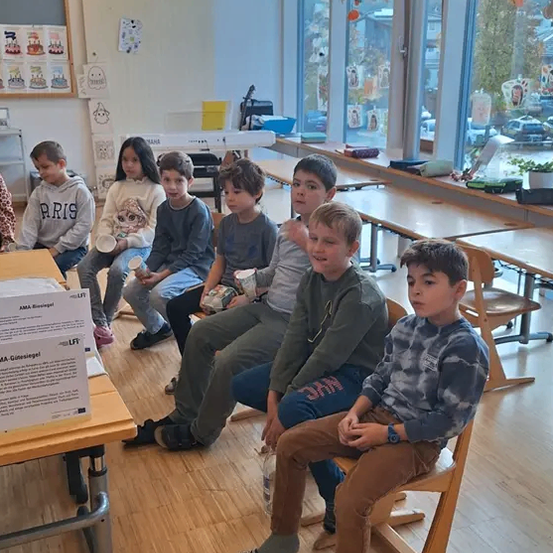 Mehrere Kinder sitzen in einem Klassenzimmer. Eines hält einen Becher, ein anderes trägt einen Schuh, und einige sitzen auf einem Holzstuhl. Eine Tafel ist an der Wand, und eine Pflanze in einem Topf befindet sich in der Nähe.