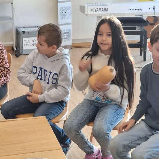 Drei Kinder sitzen in einem Klassenzimmer. Ein Mädchen hält eine Kürbis, während ein Junge mit einem Paris-Hoodie neben ihr sitzt. Ein anderer Junge sitzt rechts.