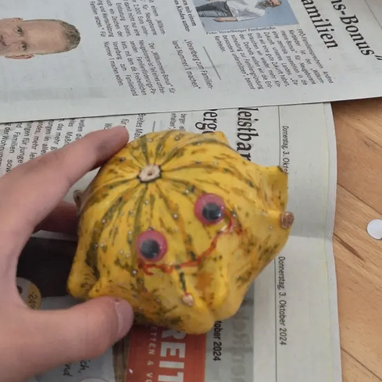 Eine Hand hält eine gelbe Keramik-Kürbisfigur mit gemalten Augen auf einer Zeitung.