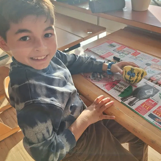 Ein junger Junge sitzt mit einer Zeitung am Tisch, hält eine bemalte Kürbis und lächelt, mit Topfpflanzen im Hintergrund.