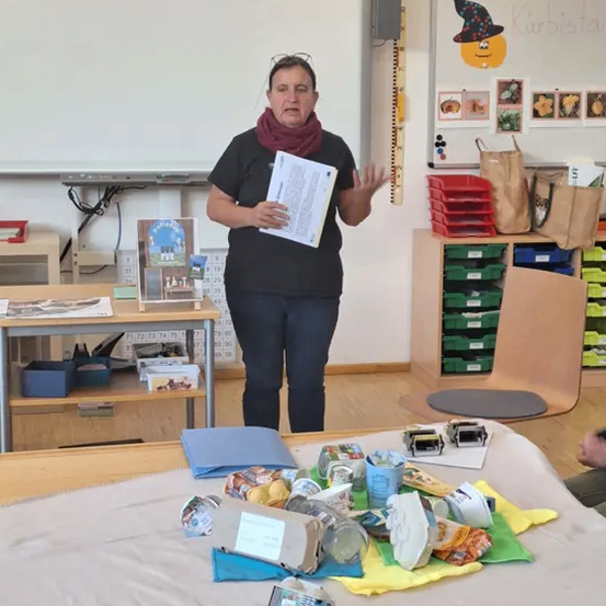 Eine Frau steht in einem Klassenzimmer neben einem Tisch voller Müll und Abfällen. Sie hält ein Papier in der Hand und spricht. Hinter ihr befindet sich eine Tafel mit Zeichnungen.