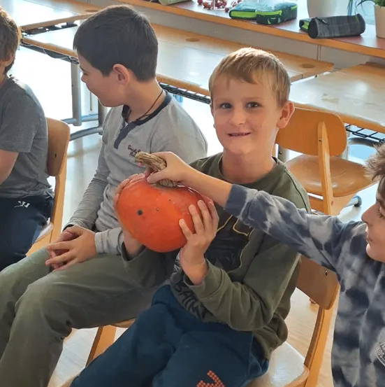 Drei Jungen sitzen in einem Klassenzimmer auf Stühlen und halten eine Kürbis. Der Junge in der Mitte trägt eine Halskette und graue Hosen. Der Junge auf der rechten Seite trägt ein grünes Langarmshirt und blaue Hosen.