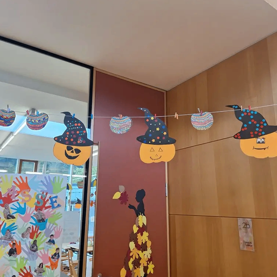 Ein Flur mit einer Wand, die mit Kürbissen, Äpfeln und einer Hexenfigur dekoriert ist. Es gibt ein farbenfrohes Poster mit Handabdrücken von Kindern.