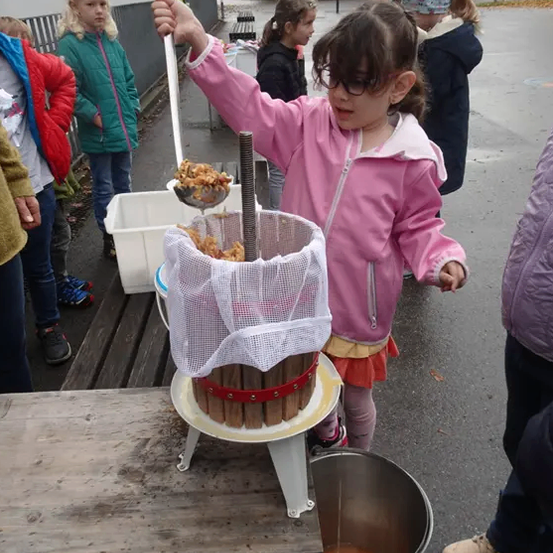 Ein Mädchen in einer rosa Jacke gießt etwas in einen Holzfass, während andere Kinder zusehen.