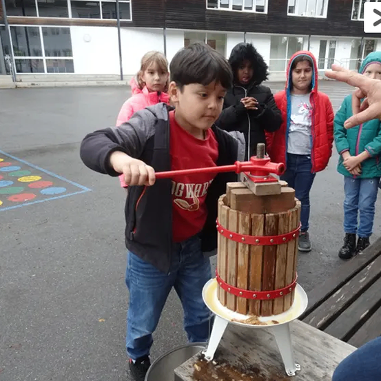Ein junger Junge in einem roten T-Shirt bedient einen hölzernen Apfelpresse im Freien. Hinter ihm beobachten drei Kinder und ein Erwachsener. Ein Gebäude ist im Hintergrund.