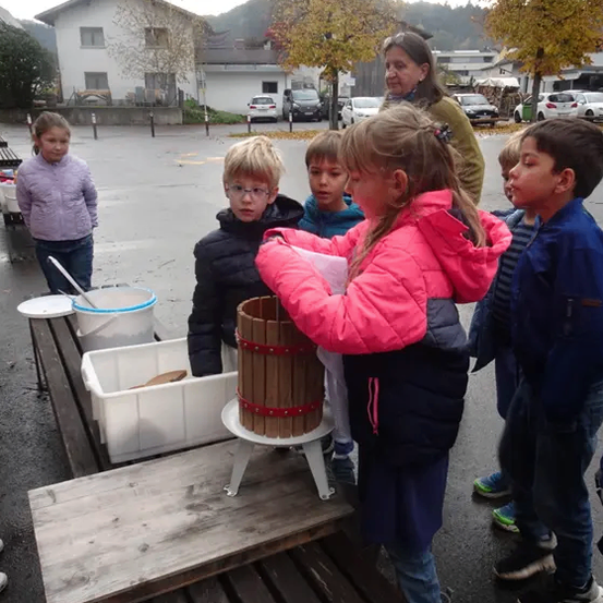 Eine Gruppe von Kindern steht um einen Holztisch herum und beobachtet, wie ein Mädchen Trauben in einem Fass presst. Eine Frau steht in der Nähe und mehrere Autos sind im Hintergrund geparkt.