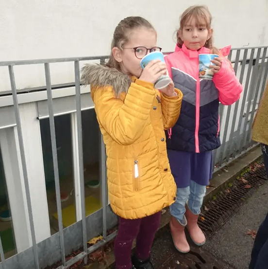Zwei junge Mädchen stehen draußen und halten jeweils einen Becher. Das Mädchen auf der linken Seite trägt eine gelbe Jacke, eine Brille und Sneakers. Das Mädchen auf der rechten Seite trägt eine rosa-graue Jacke, Stiefel und hält einen Becher. Hinter ihnen steht ein weißes Gebäude mit Glasfenstern und einem Metallzaun.