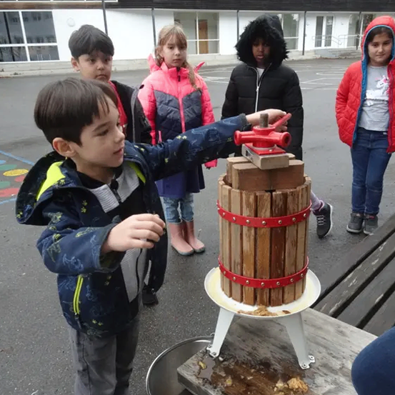 Ein junger Junge benutzt eine manuelle Apfelpresse im Freien, wobei vier Kinder hinter ihm stehen.