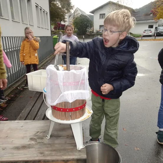 Ein junger Junge benutzt eine hölzerne Presse, um etwas aus einem Netzbeutel herauszuquetschen, mit einem Metalleimer in der Nähe, während mehrere Kinder hinter ihm stehen.