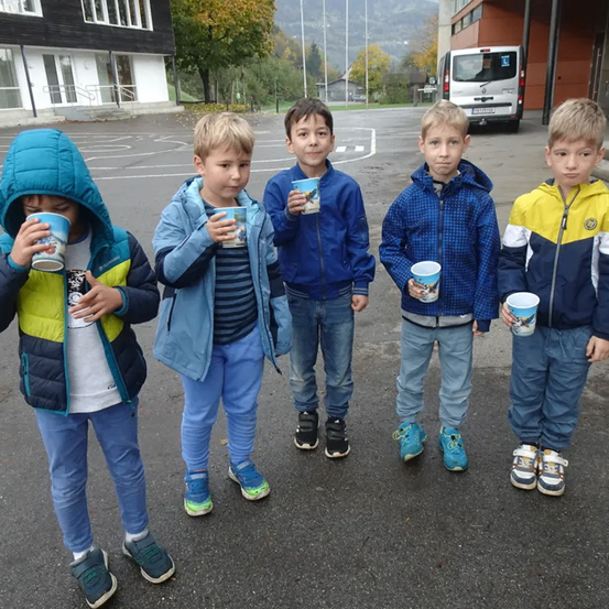 Fünf Jungen stehen auf einem Parkplatz. Jeder Junge hält einen Becher. Sie tragen Jacken und Hosen. Ein Gebäude und ein Van sind im Hintergrund.