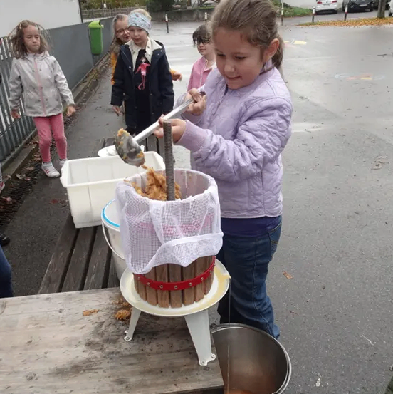Ein junges Mädchen benutzt eine hölzerne Presse, um Saft aus Früchten zu extrahieren. In der Nähe beobachten andere Kinder und einige Eimer halten den gepressten Saft.