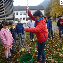 Ein Mädchen mit einer roten Jacke sammelt Bälle in einem Korb aus einem Eimer auf dem Boden vor einer Gruppe von Kindern.