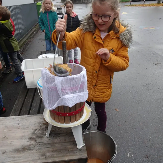 Ein Mädchen in einer gelben Jacke presst etwas durch ein Sieb in einen Eimer. Andere Kinder stehen hinter ihr.