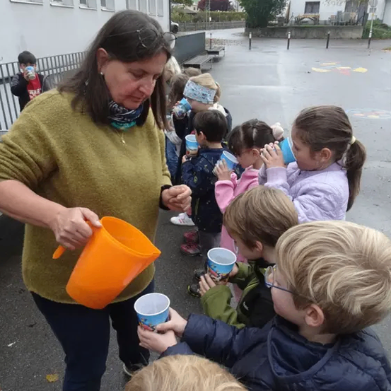 Eine Frau verteilt orangefarbene Gießkannen an eine Reihe von Kindern vor einem Gebäude. Sie halten jeweils kleine Becher.