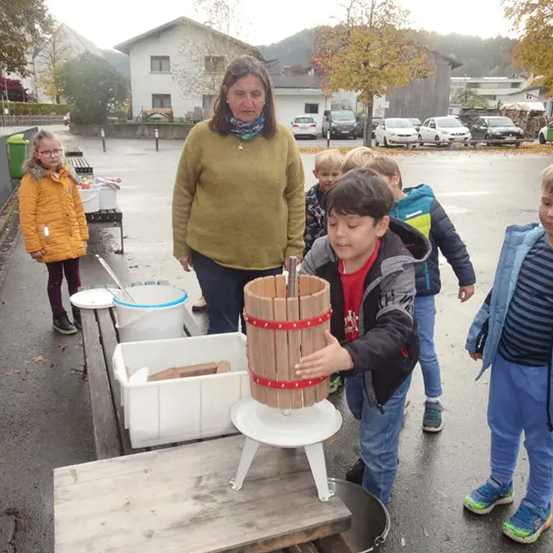 Eine Frau und mehrere Kinder sind im Freien. Die Frau steht hinter einer Holzpresse. Mehrere Kinder pressen Trauben.