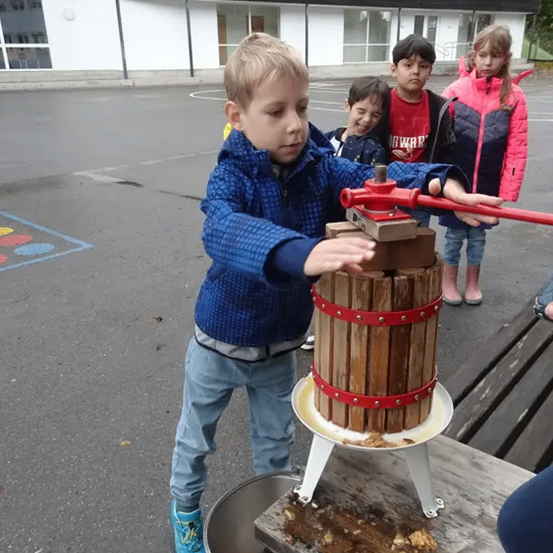 Ein junger Junge benutzt eine Holzpresse, um Saft aus Früchten zu pressen, während drei Kinder zusehen. Ein Gebäude mit Glasfenstern steht dahinter.