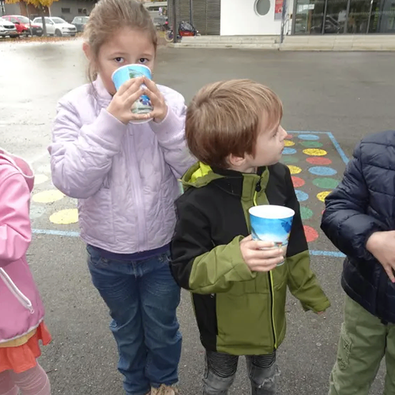 Ein Mädchen und zwei Jungen stehen auf einer nassen Straße und halten Tassen. Das Mädchen trägt eine rosa Jacke und Jeans, während die Jungen grüne und schwarze Jacken tragen. Hinter ihnen befindet sich ein Gebäude mit einem runden Fenster und einem Schild mit der Aufschrift 'Wesenbäcks'.