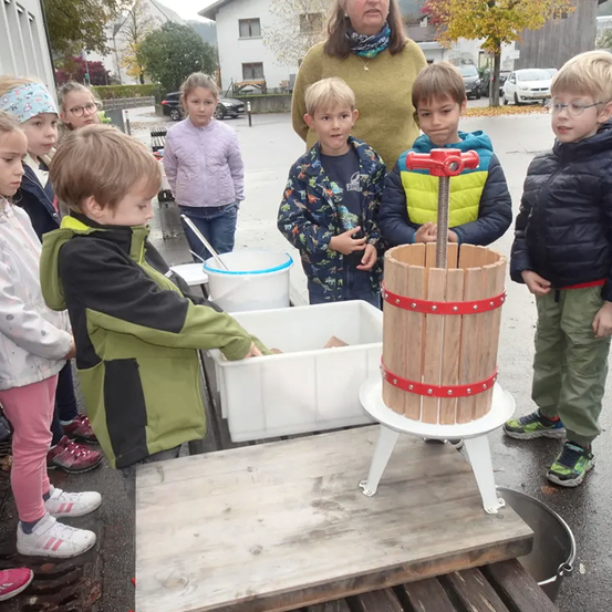 Eine Gruppe von Kindern steht um eine hölzerne Presse herum, mit einer Frau dahinter. Sie schauen einen Jungen an, der die Presse bedient. Dahinter sind Autos am Straßenrand geparkt.