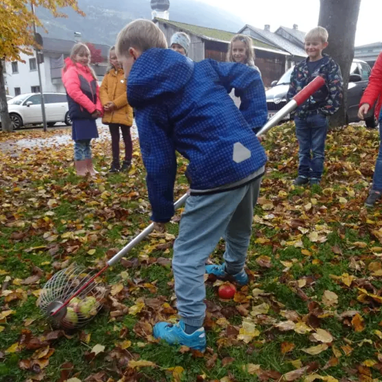 Ein junger Junge hebt Äpfel mit einem Rechen vom Boden auf, während andere Kinder zusehen.