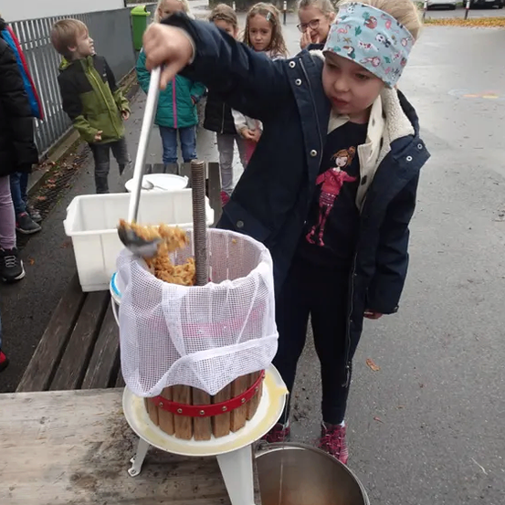 Ein kleines Mädchen mit einem Stirnband gießt Obst in eine Presse, während mehrere Kinder im Hintergrund zusehen.