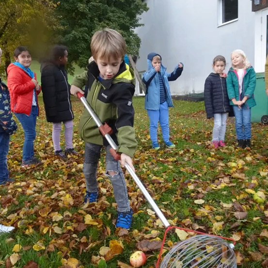 Ein Junge benutzt einen Laubbesen, um Blätter zu sammeln, während andere Kinder und Erwachsene im Hintergrund zusehen.