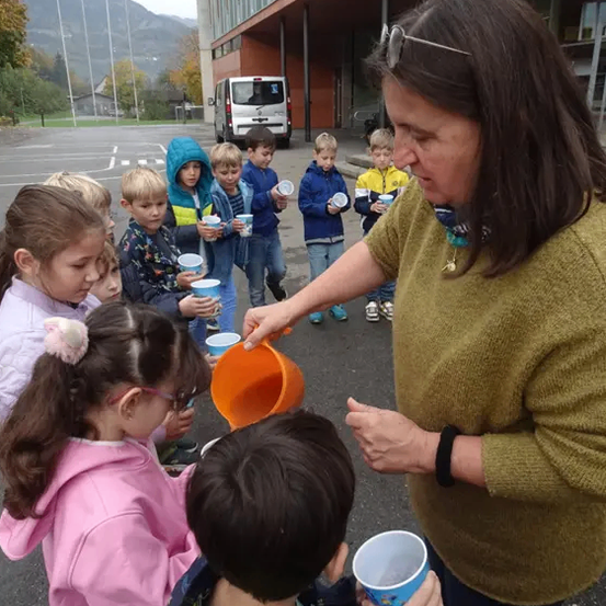 Eine Frau in einem grünen Pullover gießt orangefarbene Flüssigkeit in einen Becher, umgeben von Kindern mit Bechern, vor einem modernen Gebäude mit Bergen im Hintergrund.