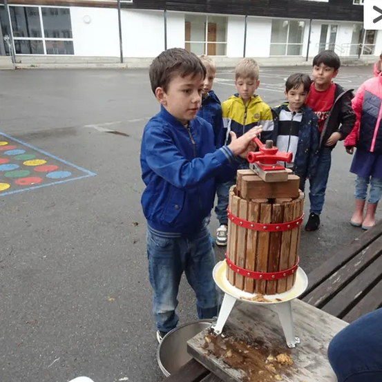 Ein junger Junge benutzt eine manuelle Presse auf dem Schulhof, während andere Kinder zusehen. Ein Gebäude mit Glasfenstern ist im Hintergrund.