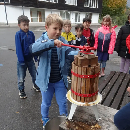 Ein junger Junge bedient eine hölzerne Presse, umgeben von Kindern, mit einem Gebäude und Bäumen im Hintergrund.