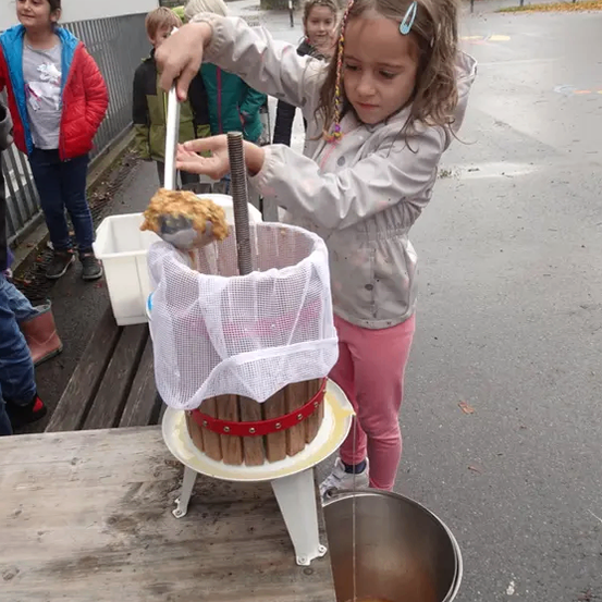 Ein kleines Mädchen benutzt eine Holzpresse, um Saft zu extrahieren, mit einem Eimer und anderen Kindern in der Nähe.