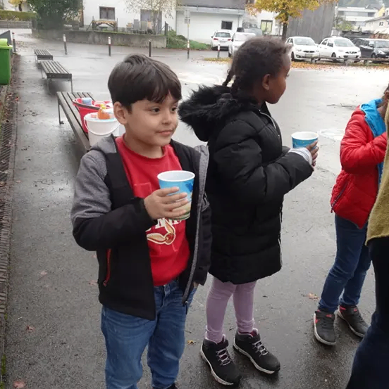 Zwei Kinder stehen auf einem nassen Parkplatz. Eines hält einen Becher und lächelt. Ein Mädchen neben ihm hält einen Becher und schaut in die Ferne. Eine Bank mit Eimern steht hinter ihnen. Autos sind in der Ferne geparkt.
