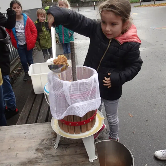 Ein junges Mädchen benutzt einen handbetriebenen Entsafter im Freien, wobei ein Eimer den Saft auffängt. Mehrere Kinder und Erwachsene stehen in der Nähe und beobachten. Der Entsafter steht auf einem Holztisch, mit einem weißen Behälter am Boden.