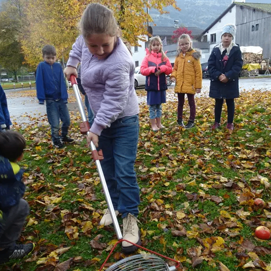 Ein Mädchen sammelt Äpfel in einem Korb auf einem Feld voller Blätter. Andere Kinder beobachten.