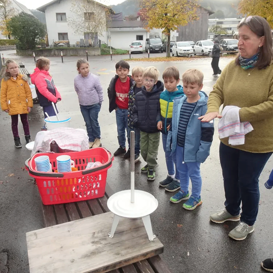 Eine Gruppe von Kindern in Jacken steht in der Nähe eines Tisches und eines roten Korbs mit Tassen. Eine erwachsene Frau steht vor ihnen.