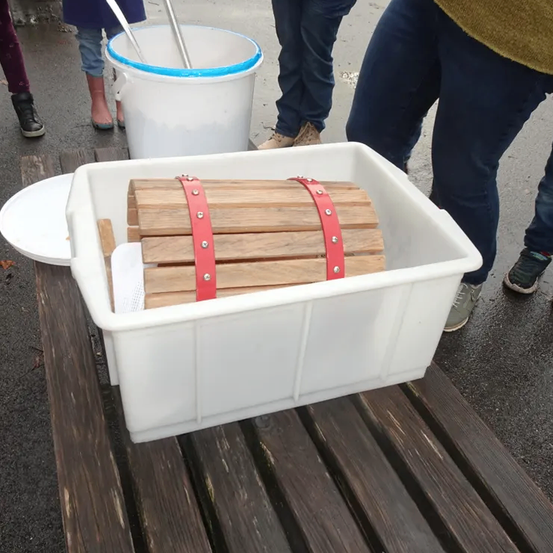 Ein hölzerner Kasten mit roten Riemen sitzt auf einer Bank draußen. Ein weißer Eimer mit blauem Rand steht in der Nähe, und mehrere Menschen sind anwesend.