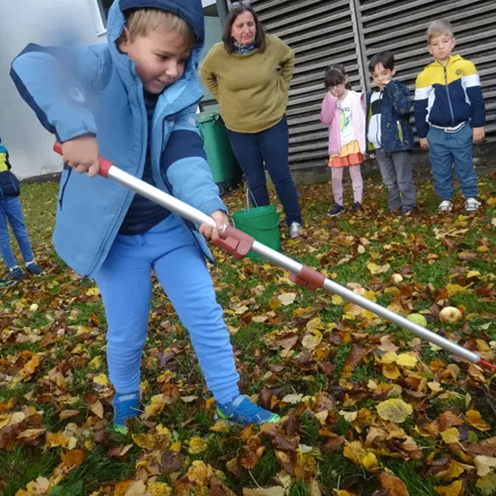 Ein Kind benutzt einen Rechen, um Blätter in einem Garten aufzuheben, während andere Kinder und Erwachsene in der Nähe sind.