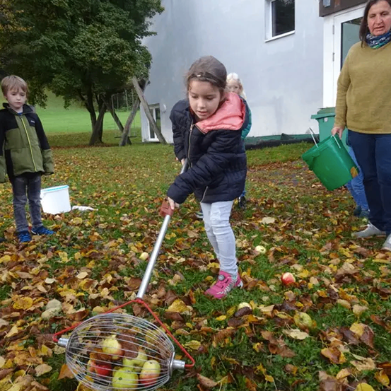 Ein junges Mädchen benutzt ein Werkzeug, um Äpfel vom Boden aufzusammeln, während ein anderes Kind und eine Frau in der Nähe stehen. Dahinter befindet sich ein Gebäude mit Balkon.