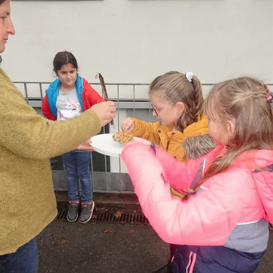 Eine Frau serviert drei Mädchen Essen auf einem Teller. Die Mädchen stehen vor einem Gebäude mit Glasfenstern.