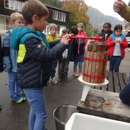 Ein junger Junge benutzt eine hölzerne Apfelpresse, umgeben von Kindern und einem Gebäude im Hintergrund.