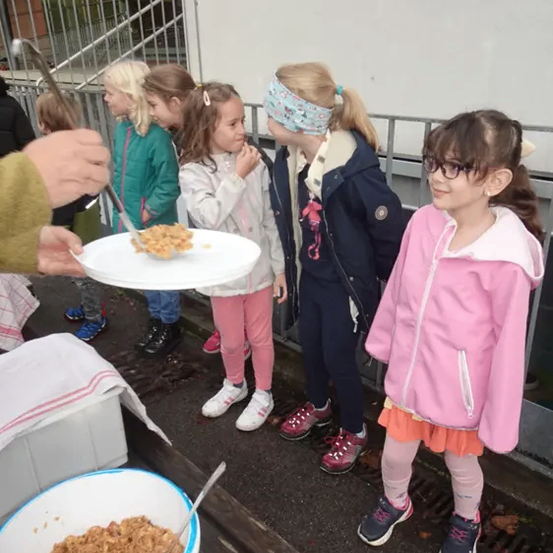 Eine Gruppe von Kindern steht draußen in der Nähe eines Gebäudes, eine Person füttert sie von einem Teller und ein Eimer mit Essen steht auf dem Boden.