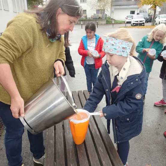 Eine Frau gießt Orangensaft in die Tasse eines Kindes auf einer Bank, mit anderen Kindern und Erwachsenen im Hintergrund.