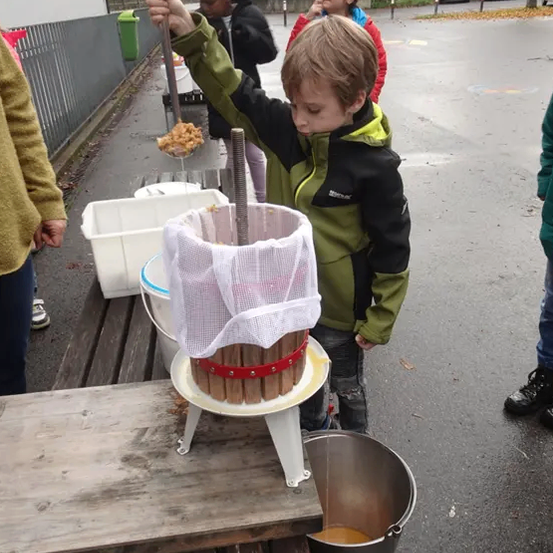 Ein junger Junge benutzt draußen eine handbetriebene Apfelpresse, während andere in der Nähe stehen. Er presst Äpfel in einen Metalleimer, während ein anderer Eimer den Saft sammelt.