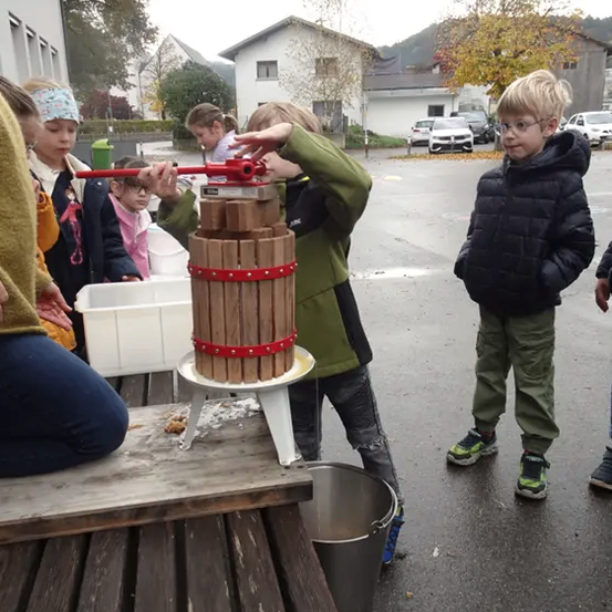 Eine Gruppe von Kindern, einschließlich eines Jungen mit Brille, ist draußen und benutzt eine manuelle Apfelpresse, um Saft zu extrahieren. In der Nähe befinden sich Eimer und ein Fass.