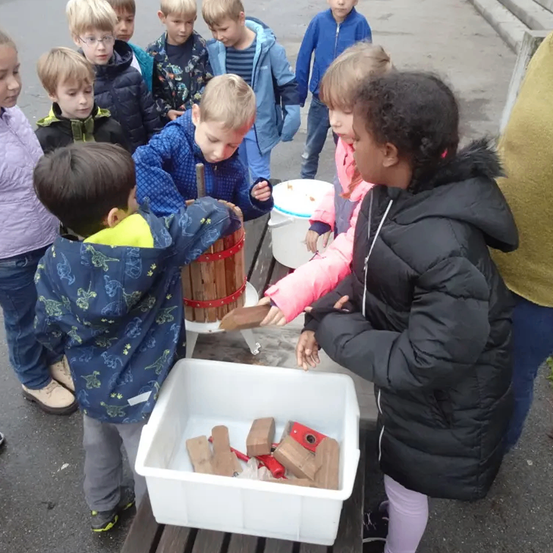 Eine Gruppe von Kindern steht draußen um eine hölzerne Presse herum, wobei ein Kind sie bedient. In der Nähe befindet sich ein weißer Plastikbehälter, der mit Holzblöcken gefüllt ist.