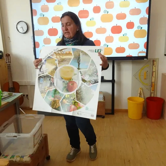 Eine Frau steht in einem Klassenzimmer und hält eine runde Tafel mit Bildern von Äpfeln und Kürbissen. Sie trägt ein schwarzes Shirt und Hose. Hinter ihr steht ein Monitor, der Kürbisse anzeigt.