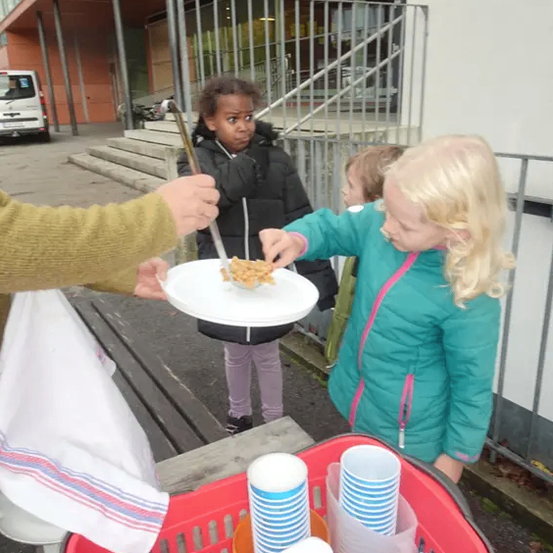 Ein Kind hält einen Teller mit Essen in der Hand und ein anderes Kind streckt danach. Es gibt einen roten Korb mit Bechern und einem Handtuch auf dem Boden.