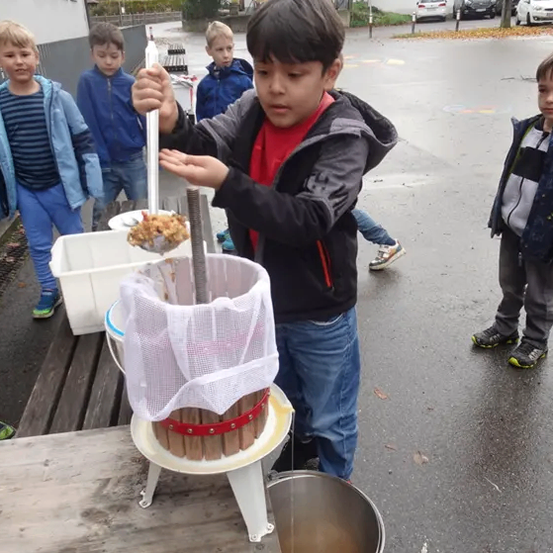 Ein Junge benutzt eine manuelle Presse, um Saft aus Früchten zu extrahieren. Mehrere Kinder beobachten. Die Einrichtung steht auf einem Holztisch mit einem Eimer darunter.