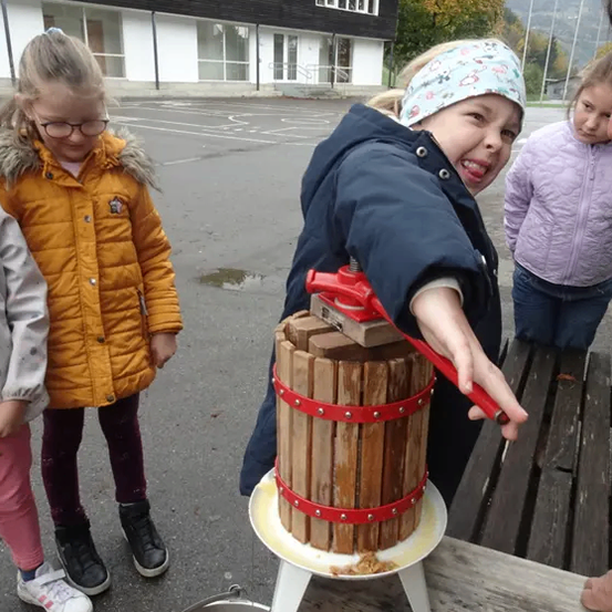 Drei junge Mädchen auf einem Schulhof benutzen eine Holzpresse. Das Mädchen in der Mitte lächelt, während sie die Presse zusammendrückt. Dahinter steht ein Gebäude und ein Baum.