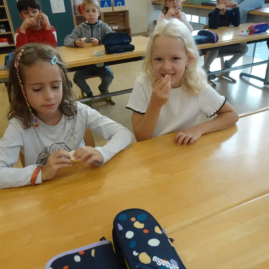 Mehrere Kinder sitzen an Tischen in einem Klassenzimmer. Zwei Mädchen, eines mit geflochtenen Haaren und eines mit blonden Haaren, essen. Verschiedene Schulutensilien liegen auf den Tischen.
